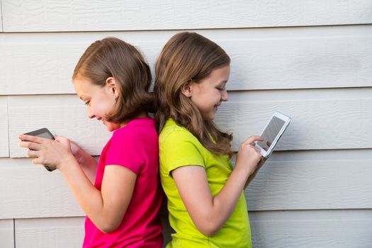 Twin sister girls playing with tablet pc happy on white wall leaning on her back