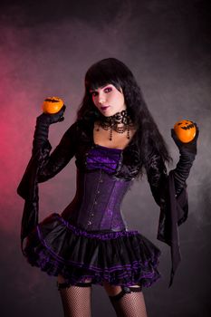 Attractive witch in purple gothic Halloween costume holding Jack-o-lantern style oranges 