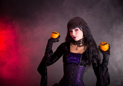 Portrait of attractive witch in purple gothic Halloween costume holding Jack-o-lantern style oranges 