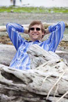 Handsome Caucasian man in forties leaning back against log on beach, relaxed with arms behind head.