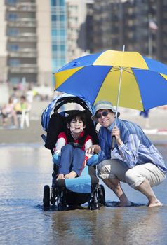 Father holding umbrella over disabled son in wheelchair at the beach