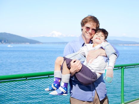Father holding disabled son in arms on deck of ferry boat. Puget Sound in background. Child has cerebral palsy.