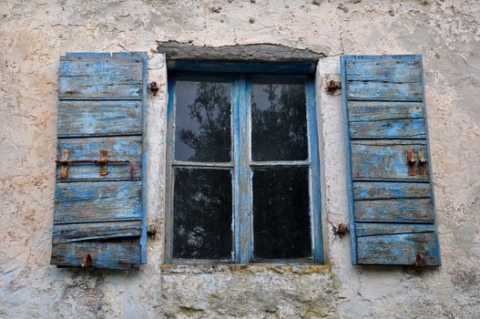 Wooden window shutter with chipped blue paint and textured wall of old house.