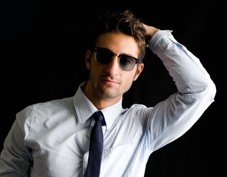 Handsome, elegant young man with shirt, tie and sunglasses, one hand on his head, isolated on black