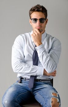 Handsome young man wearing shirt, necktie and ripped jeans, sitting, isolated on grey background