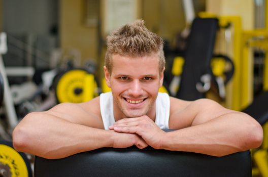 Handsome young man in gym resting on gym equipment, smiling and looking in camera