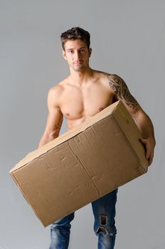 Handsome, athletic, shirtless young man carrying big cardboard box, isolated on grey background