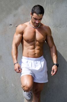 Muscular male bodybuilder standing against wall outdoors shirtless, wearing white boxer shorts