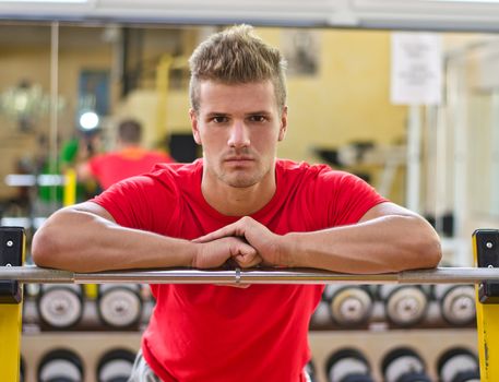 Handsome young man in gym resting on barbell, looking in camera