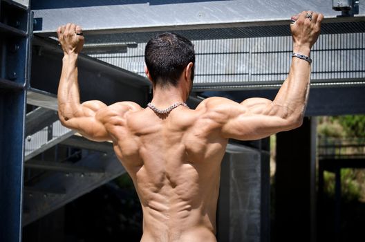 Muscular back of shirtless male bodybuilder hanging from metal structure outdoors