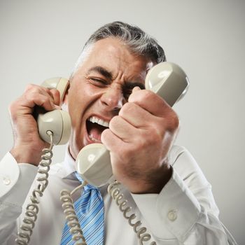 A stressed businessman yells loudly into the three handsets that he is holding.