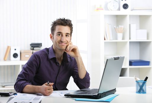 Young businessman working on laptop, looking at camera