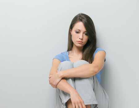 Young sad woman sitting on floor against grey background