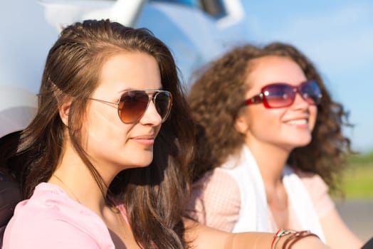 Two attractive young women wearing sunglasses, sitting next to the car