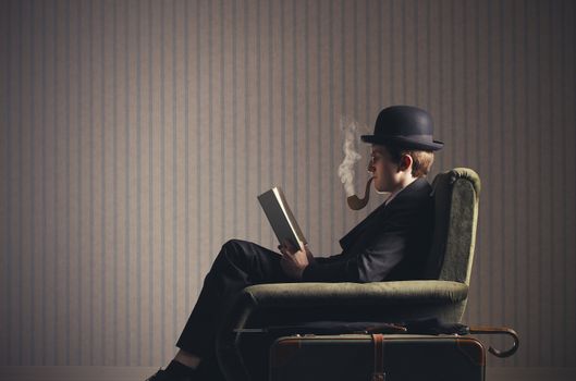 Bizarre man sitting on armchair reading a book and smoking a fake pipe 