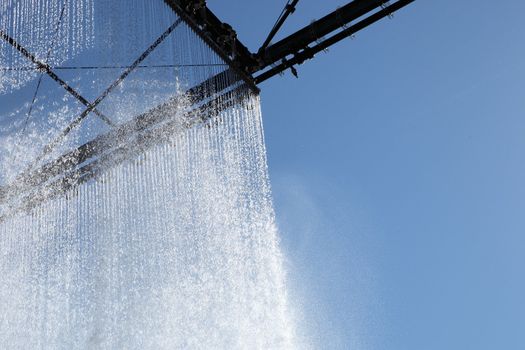 Sparkling ornamental curtain of falling water catching the sunlight as it falls from an overhead structure against a blue summer sky with copyspace