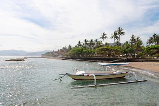 Traditional wooden fishing boat anchored near a beach with green palms and blue sky on background. Bali, Indonesia. 