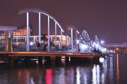 night image of scenic pedestrian walkway in Barcelona Port Vell area with walking people