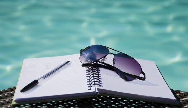 Sunglasses and pen on a note-pad  by the swimming pool