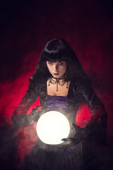 Beautiful gothic style fortune teller with a crystal ball, studio shot over smoky background 