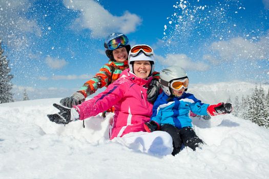 Skiing, winter, family - smiling boy in ski goggles and a helmet with his mother and sister playing in snow in winter resort