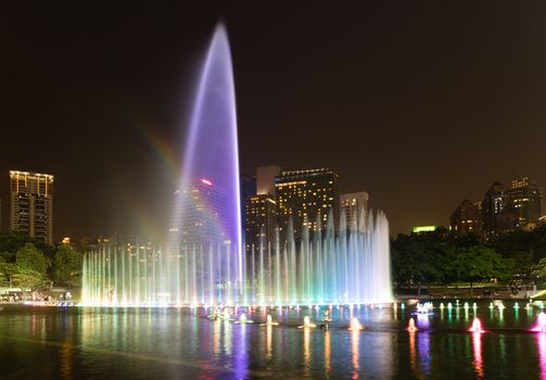 Illuminated fountain with rainbow at night in modern city skyline