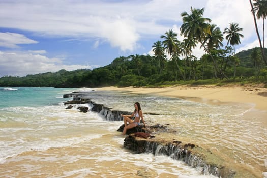 Young woman in bikini sitting on rocks at Rincon beach, Samana peninsula, Dominican Republic