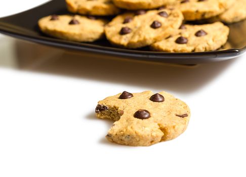 Closeup of chocolate chip cookie with a bite and pile of cookies in a square black plate on white background