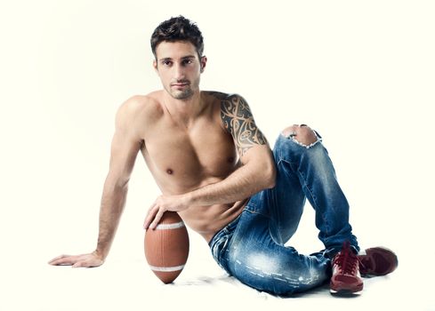 Athletic young man shirtless wearing jeans, with american football ball sitting on floor, isolated