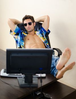 Attractive, fit young man with his shirt open sitting at office desk with feet up on it, thinking about vacation