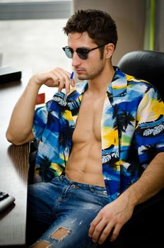 Attractive, fit young man with his shirt open sitting at office desk barechested thinking about vacation