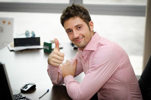 Cheerful office worker at desk doing thumb up sign and smiling. Real workplace