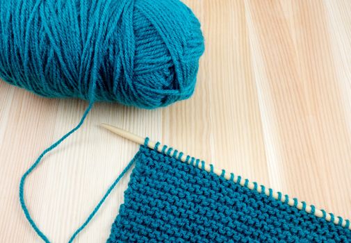Closeup of ball of teal wool with length of garter stitch on a knitting needle, on wood