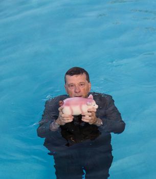 Senior caucasian man holding piggy bank above water as he slowly drowns in debt wearing business suit