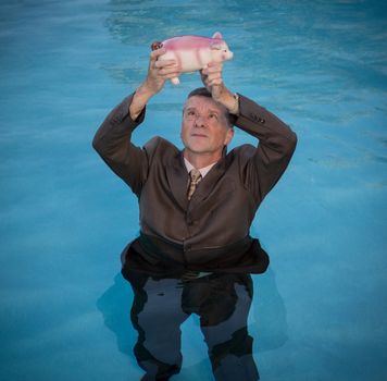 Senior caucasian man holding piggy bank above water as he slowly drowns in debt wearing business suit