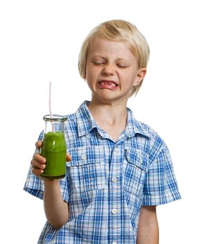 A cute funny boy is pulling a face looking at a green smoothie or juice. Isolated on white.