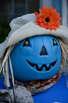 A blue plastic pumpkin serves as a scarecrow's head wearing a hat, scarf, and blue shirt