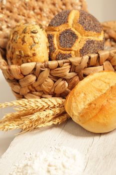 Assorted bread rolls with ears of wheat displayed in a woven straw basket on a rustic white wooden board with copyspace, close up view