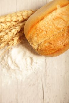 Crusty golden freshly baked roll displayed on a rustic white painted wooden board with ears of ripe wheat and flour
