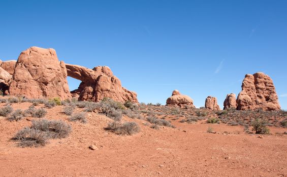 This image shows an arch and formation up a slope with a blue sky background. Taken at Arches National Park.