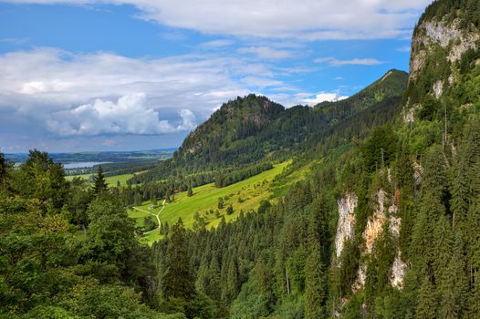 View of sunlit green meadow surrounded by forest-covered mountains under beautiful cloudy sky in Bavaria, Germany.