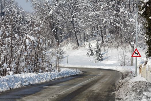 Gray curved road through trees and hills covered by snow at winter in Piedmont, Northern Italy.