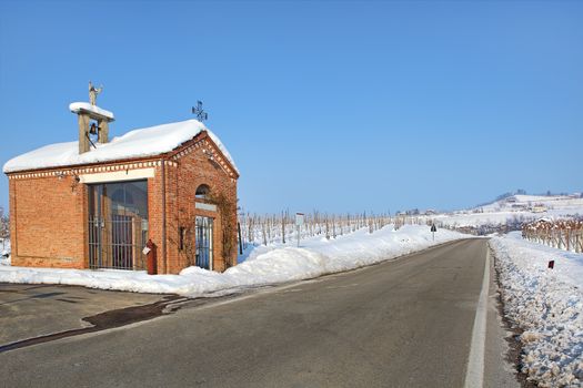 Small red chapel on the roadside among vineyards covered by white snow under clear blue winter sky in Piedmont, Northern Italy.