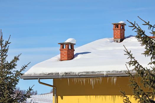 Two red brick chimneys and row of icicles on the roof covered by snow under blue winter sky in Piedmont, Northern Italy.