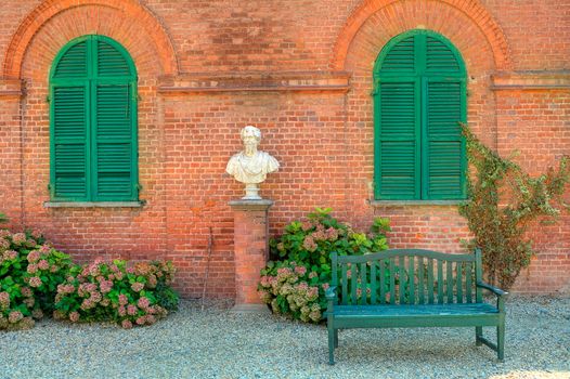 Wooden bench stand on the gravel in front of red brick house with closed green shutters in park of Racconigi in Piedmont, Northern Italy.