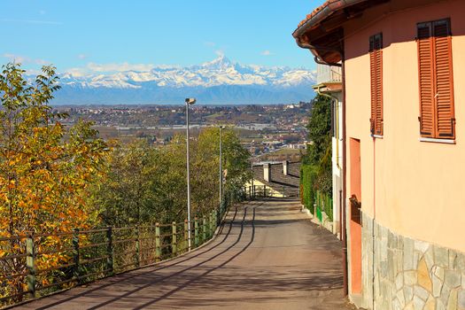 Narrow street runs through small typical italian town of Santa Vittoria D'Alba as snowy mountain peaks on background in Piedmont, Northern Italy.