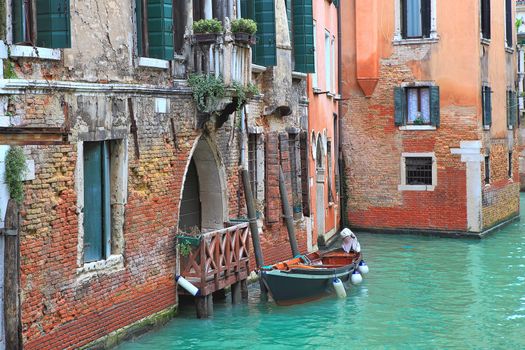 Small boat tied next to old red brick house with wooden balcony on narrow canal in Venice, Italy.