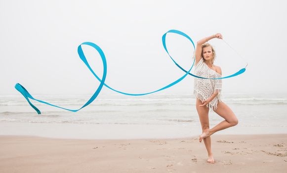 Young beautiful gymnast woman dance with ribbon on the beach at foggy day