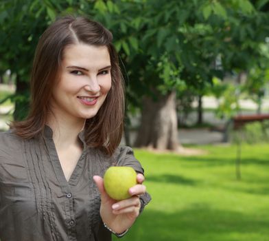 Young smiling woman offering a green apple outside in a park in summer.