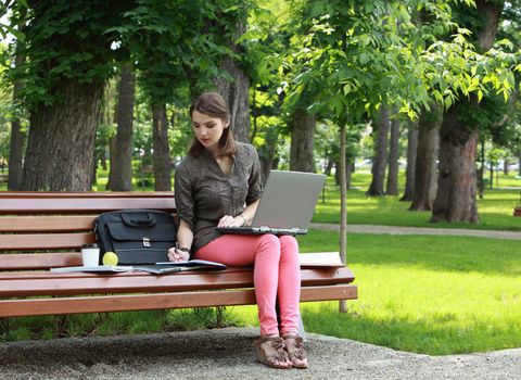 Young woman with a laptop writing something on a notebook while studying outside in a park.